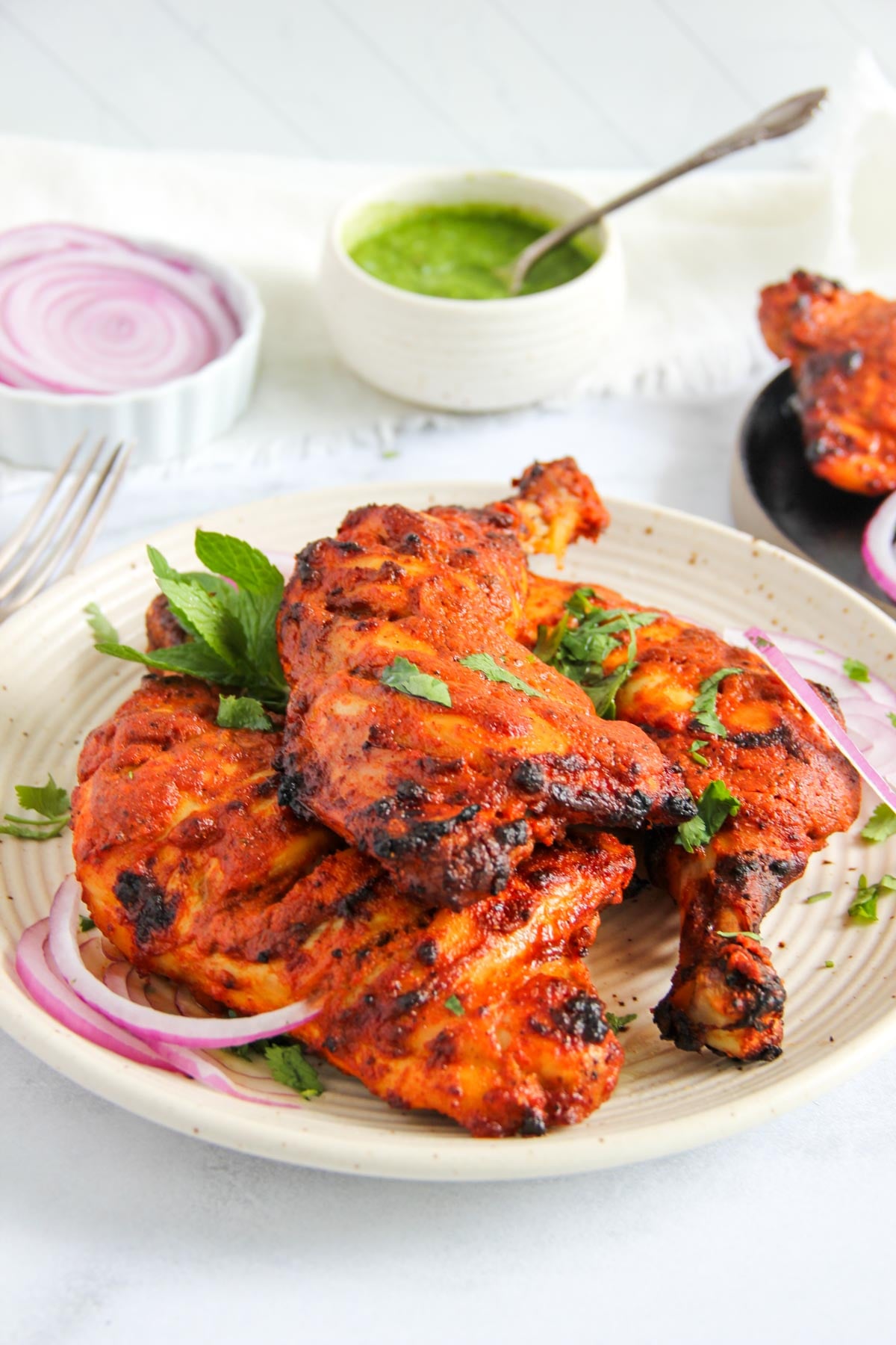 A plate of grilled tandoori chicken garnished with fresh herbs and sliced red onions, with a bowl of green chutney and additional onions in the background.