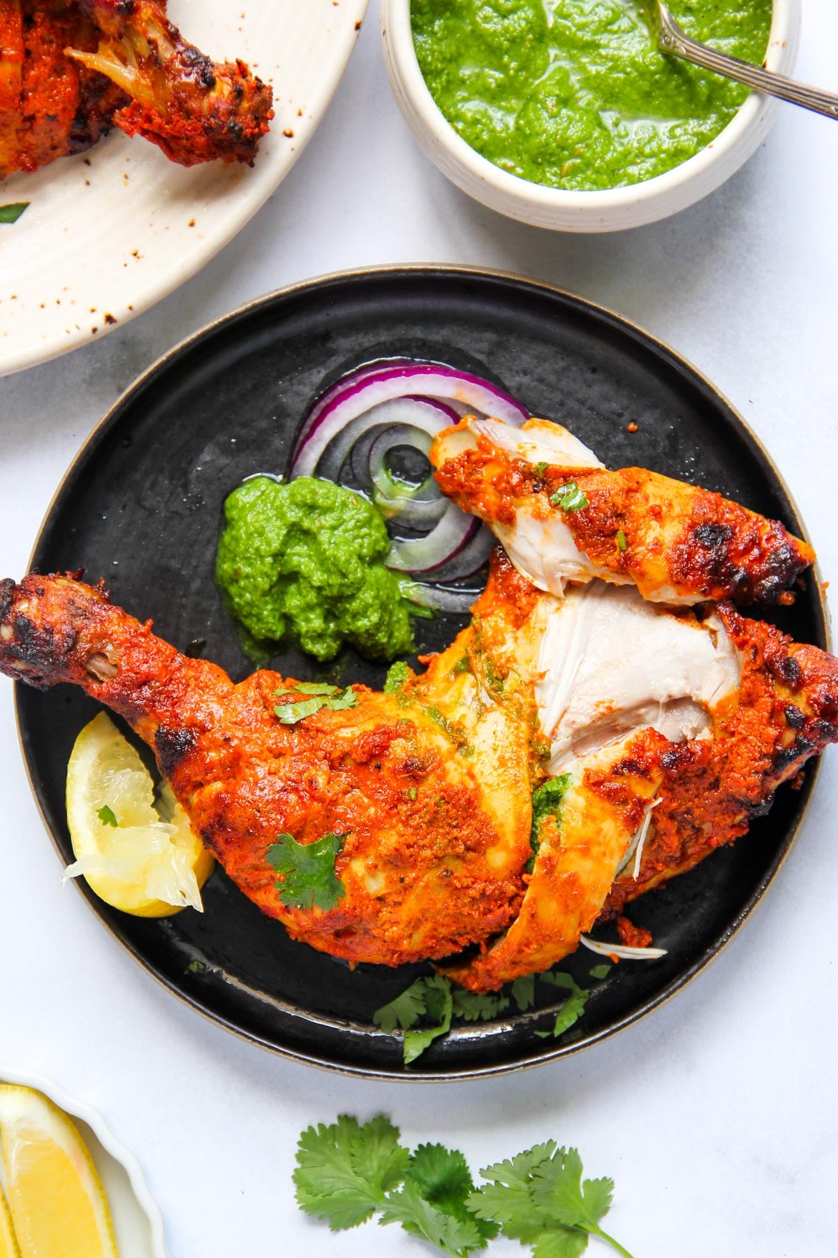 A black plate with a serving of tandoori chicken, sliced red onion, green chutney, a lemon wedge, and cilantro garnish.