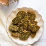 A plate of Maharashtrian kothimbir vadi garnished with sesame seeds is displayed on a white surface next to a glass of tea.