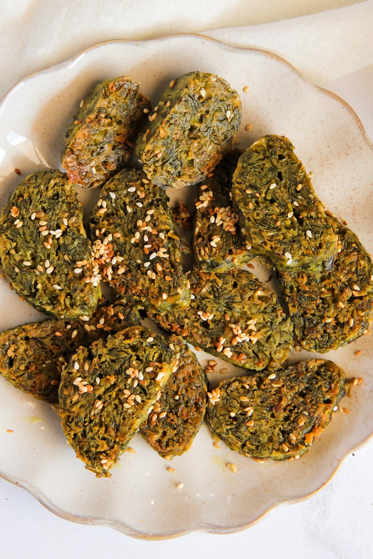 A plate of sliced green vegetable patties topped with sesame seeds, arranged on a light-colored ceramic dish.