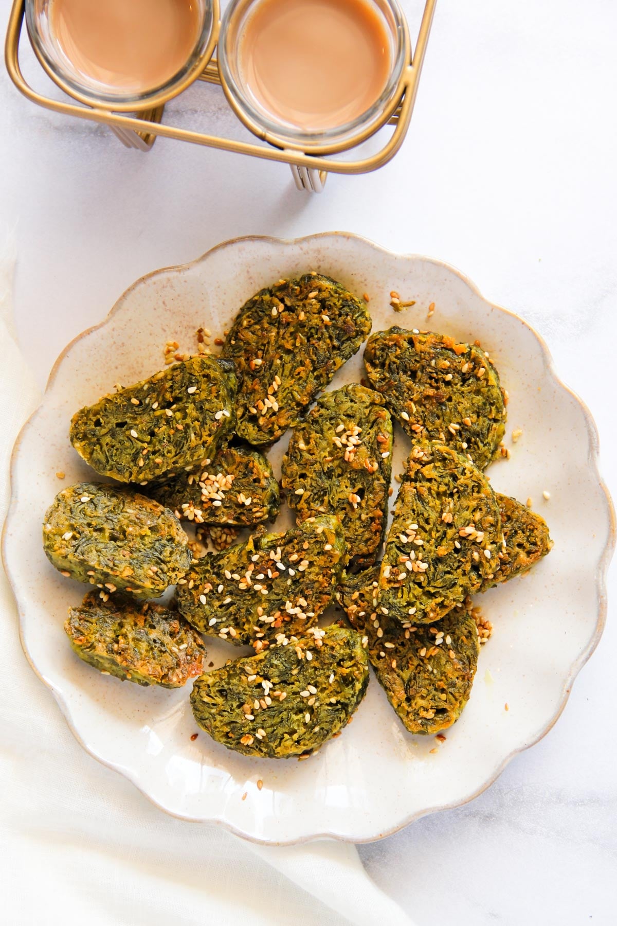 A plate of sliced, greenish-brown savory snacks garnished with sesame seeds, with two glasses of chai tea in the background.