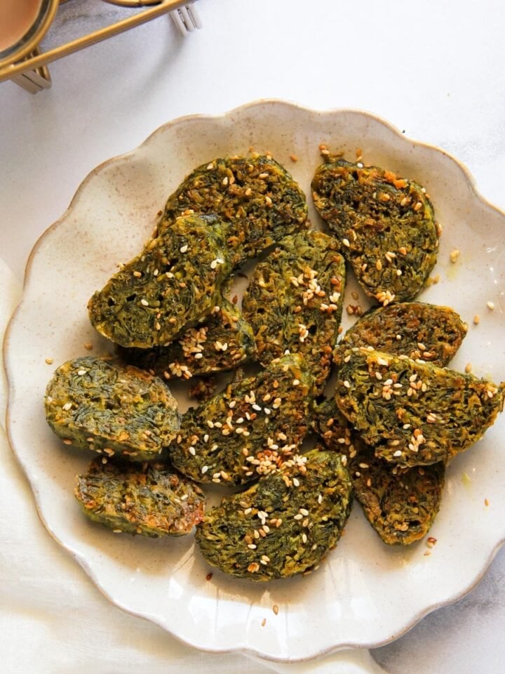 A plate of Maharashtrian kothimbir vadi garnished with sesame seeds is displayed on a white surface next to a glass of tea.