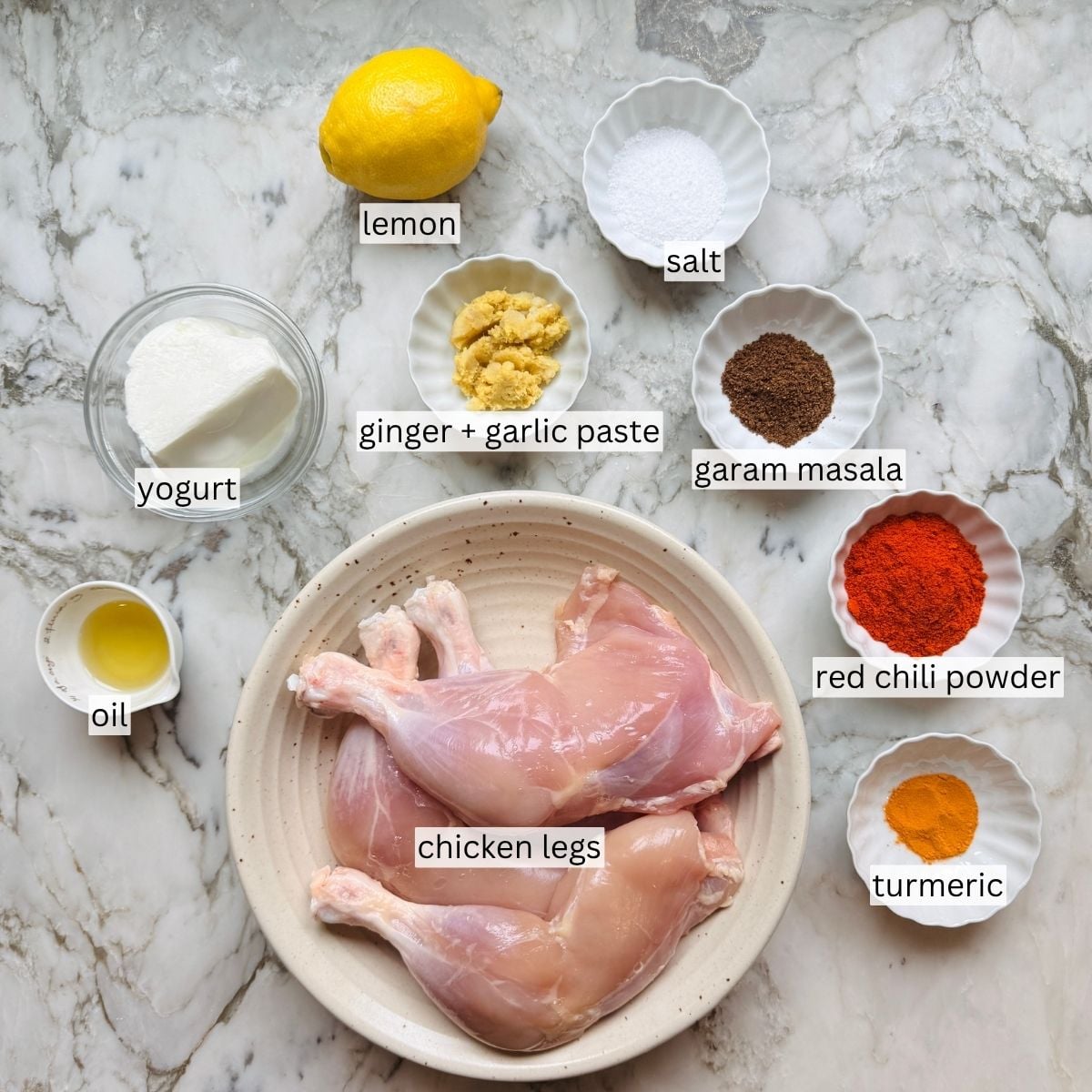 A bowl with raw chicken legs and small bowls of yogurt, lemon, oil, ginger-garlic paste, salt, garam masala, red chili powder, and turmeric on a marble surface.