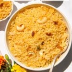 A bowl of Narali Bhat (sweet coconut rice) garnished with cashews and raisins, placed on a white surface with a spoon and colorful flowers nearby.