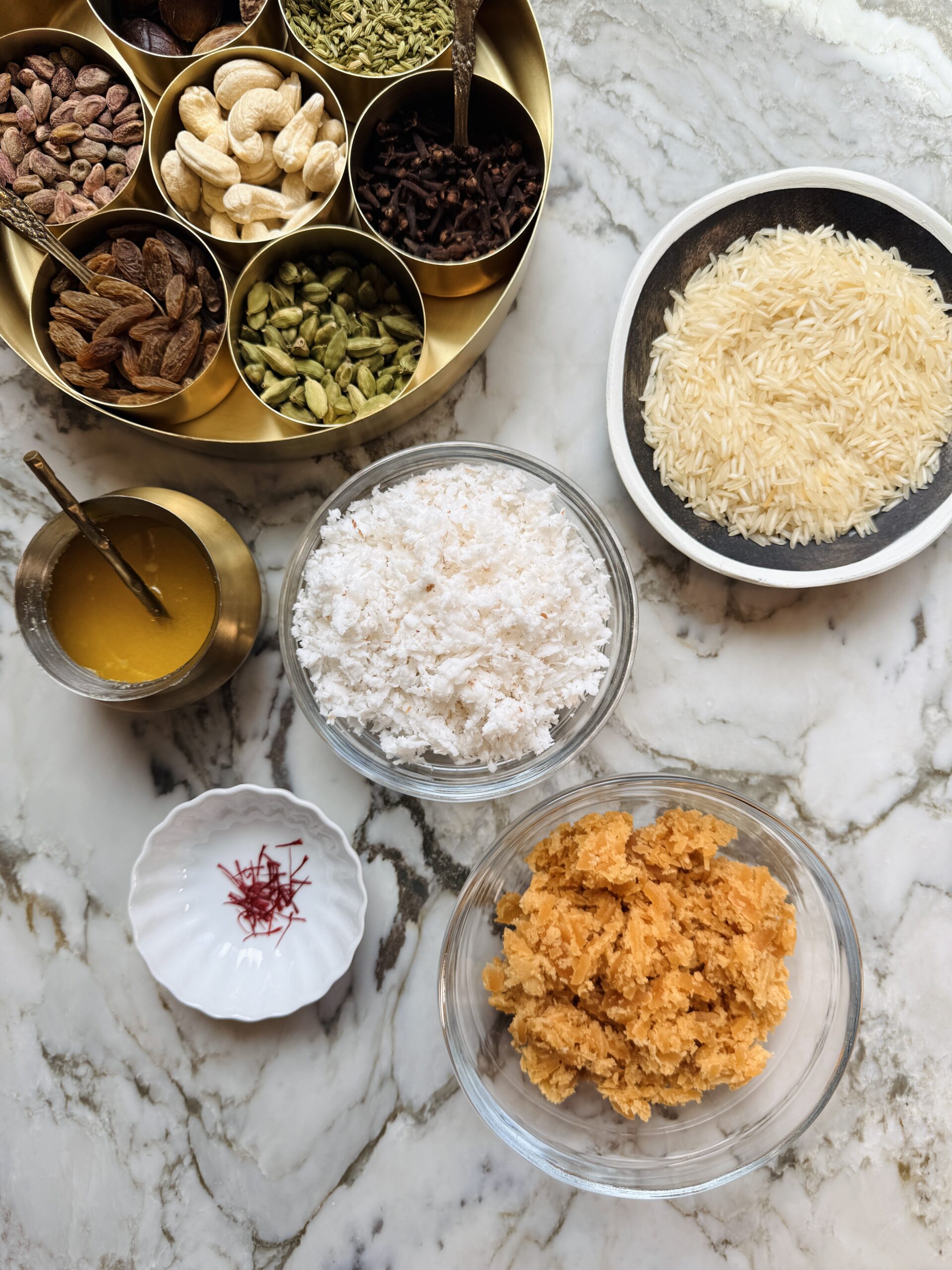 A marble table with bowls of dry fruits, rice, shredded coconut, saffron strands, ghee, cardamom pods, and grated jaggery arranged neatly.