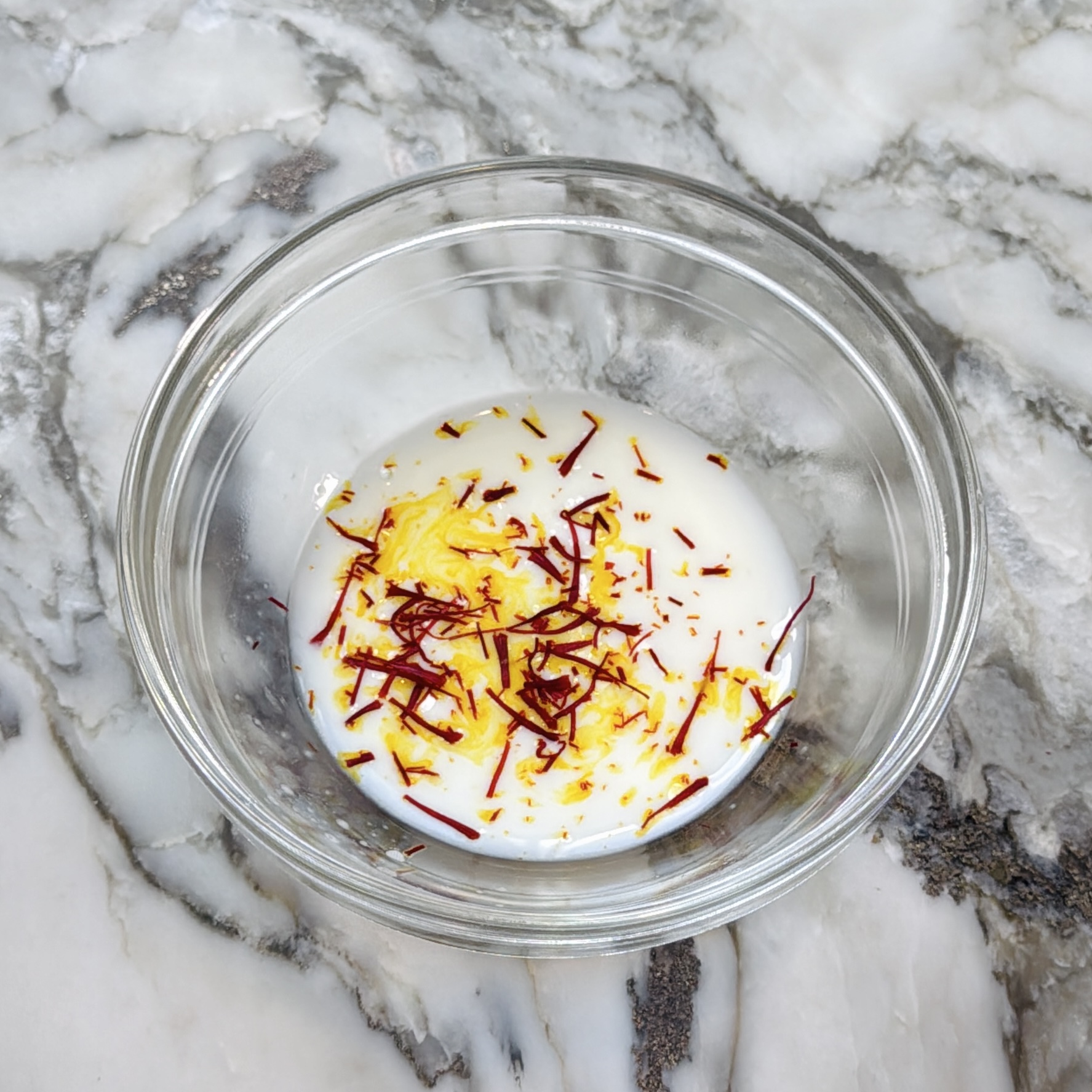 A glass bowl on a marble surface contains milk with saffron strands soaking in it.