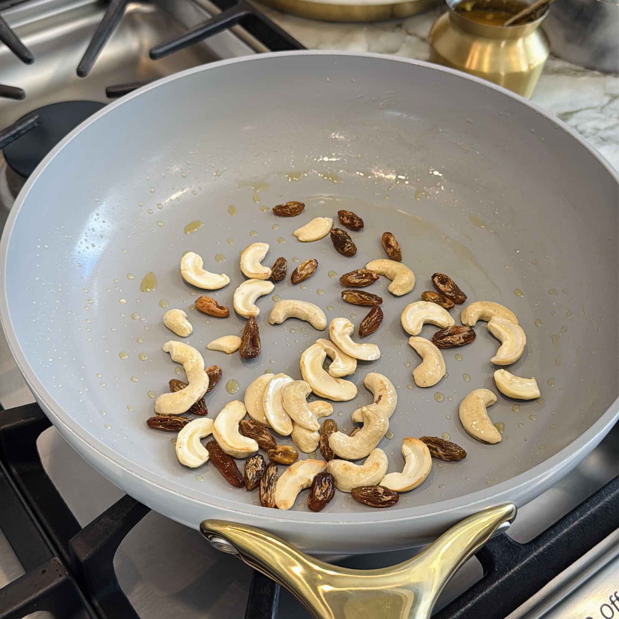 Cashews and raisins are being sautéed in a light-colored nonstick pan on a stovetop with some oil.
