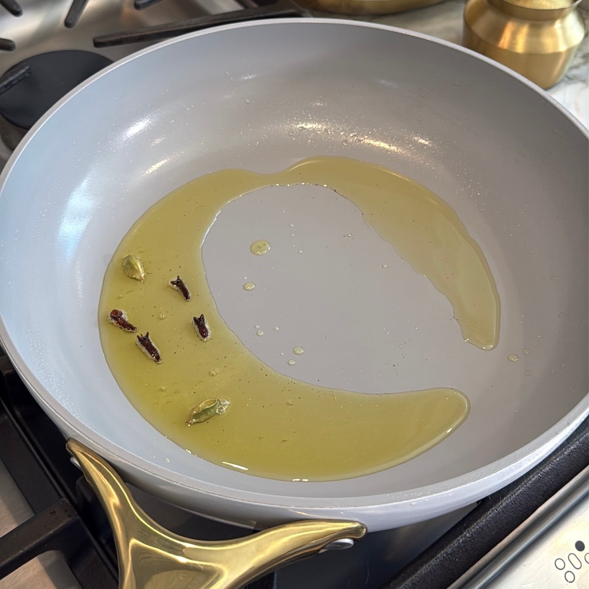 A grey frying pan with hot oil, three cloves, and two green cardamom pods sits on a stovetop, ready for cooking.