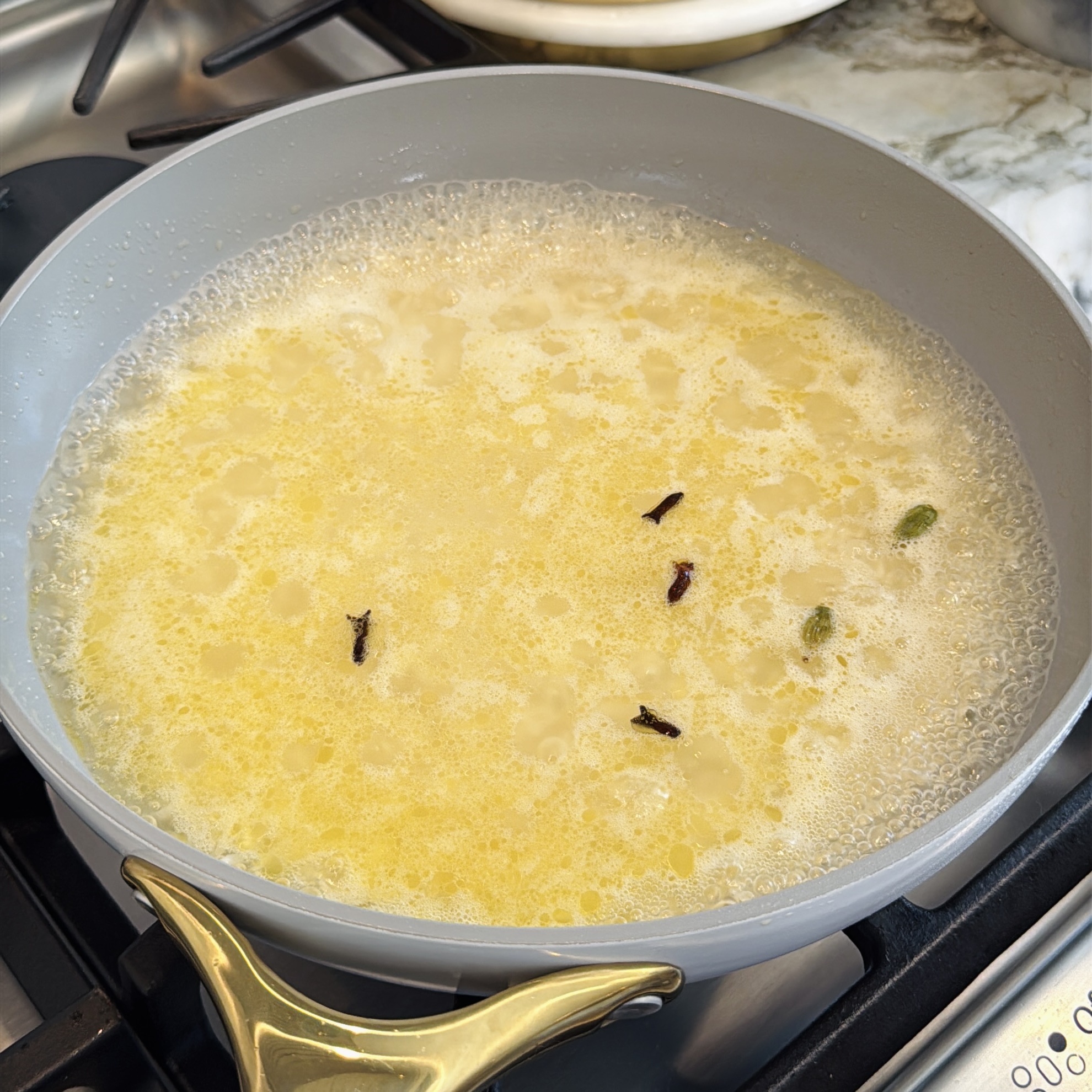 A gray pan on a stove contains boiling yellow liquid with visible oil, cloves, and a bay leaf in it.