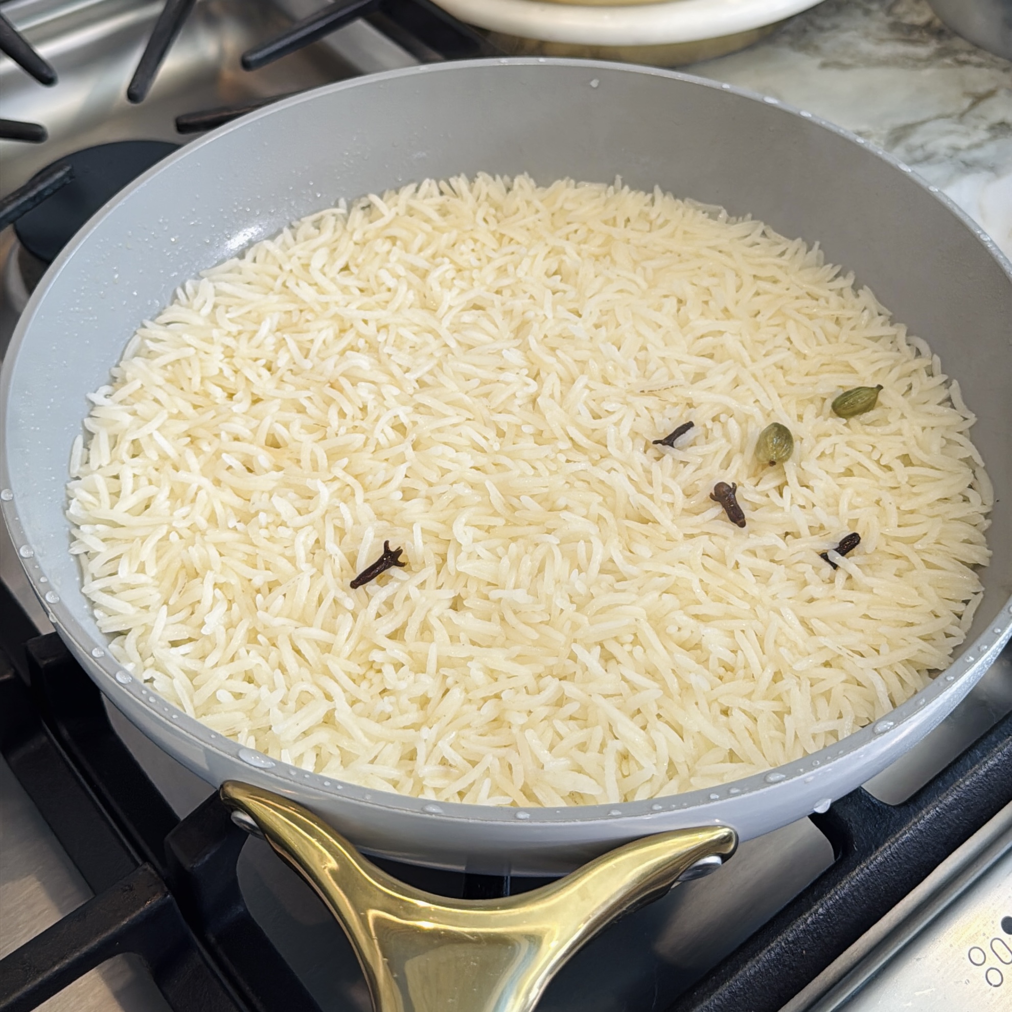 A shallow pan of white basmati rice cooking on a stove with visible whole spices, including cloves and cardamom pods, in the water.
