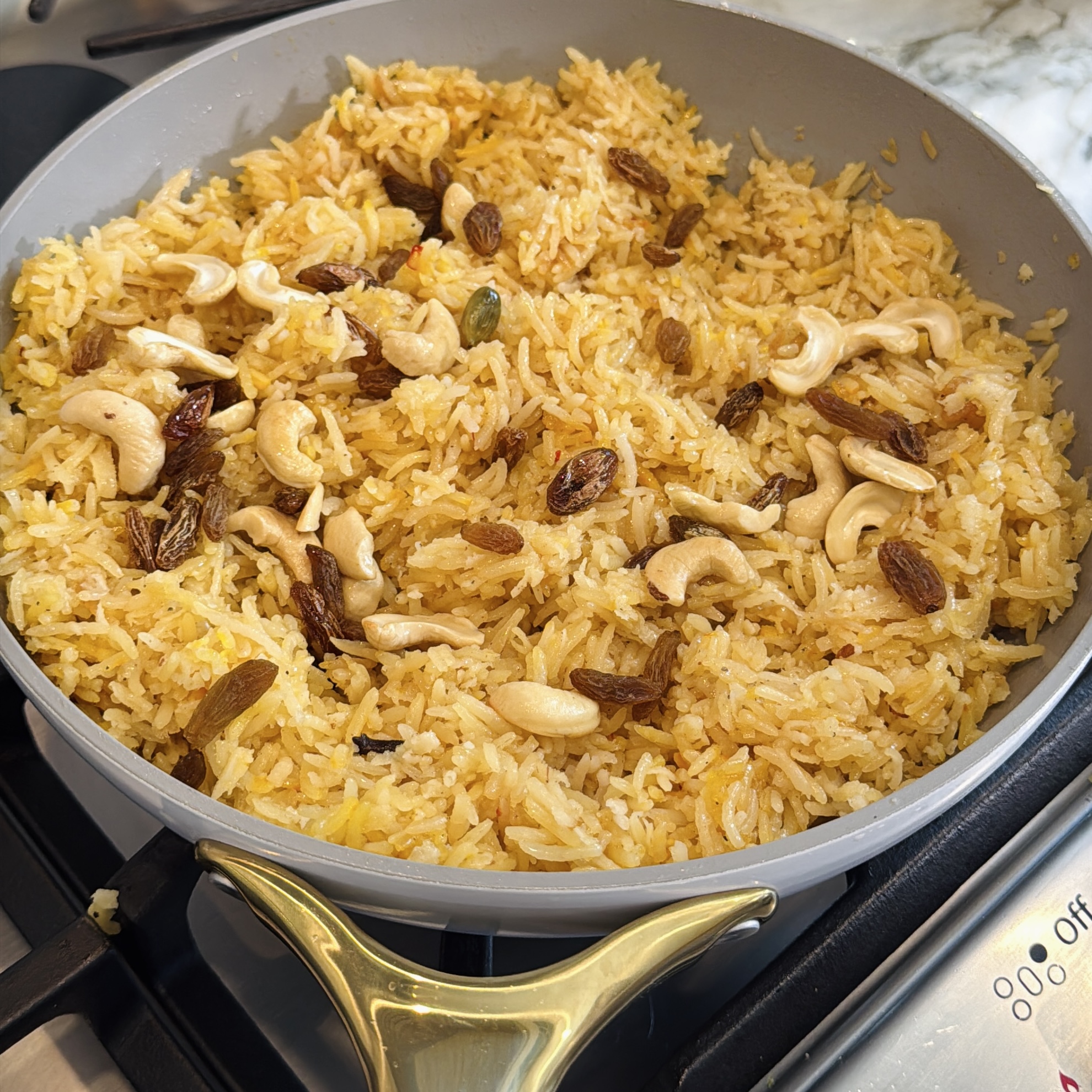 A pan filled with cooked yellow rice topped with cashews and raisins sits on a stovetop.