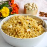 A bowl of sweet coconut rice garnished with cashews and raisins, with a spoon inside. Flowers and a small bowl of cashews are in the background.