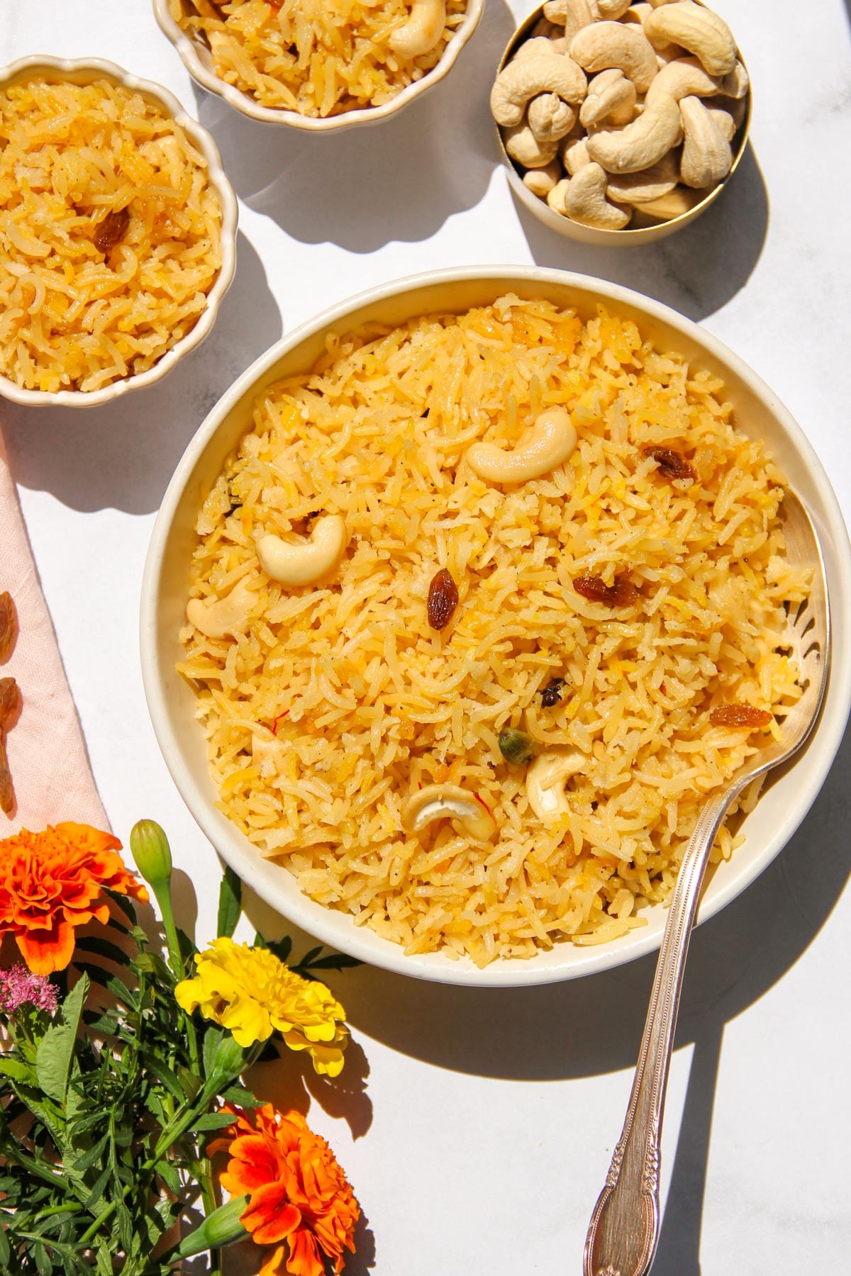 A bowl of narali bhat garnished with cashews and raisins, surrounded by smaller bowls of rice, cashews, and flowers on a white surface.