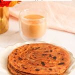 A plate of stacked pumpkin parathas sits next to a glass of chai on a marble surface, with a vase of orange flowers in the background.