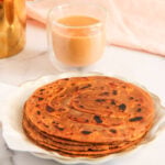 A plate of stacked parathas sits next to a glass of chai on a white surface with a pink cloth in the background.