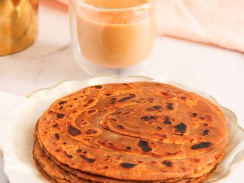 A plate of stacked parathas sits next to a glass of chai on a white surface with a pink cloth in the background.