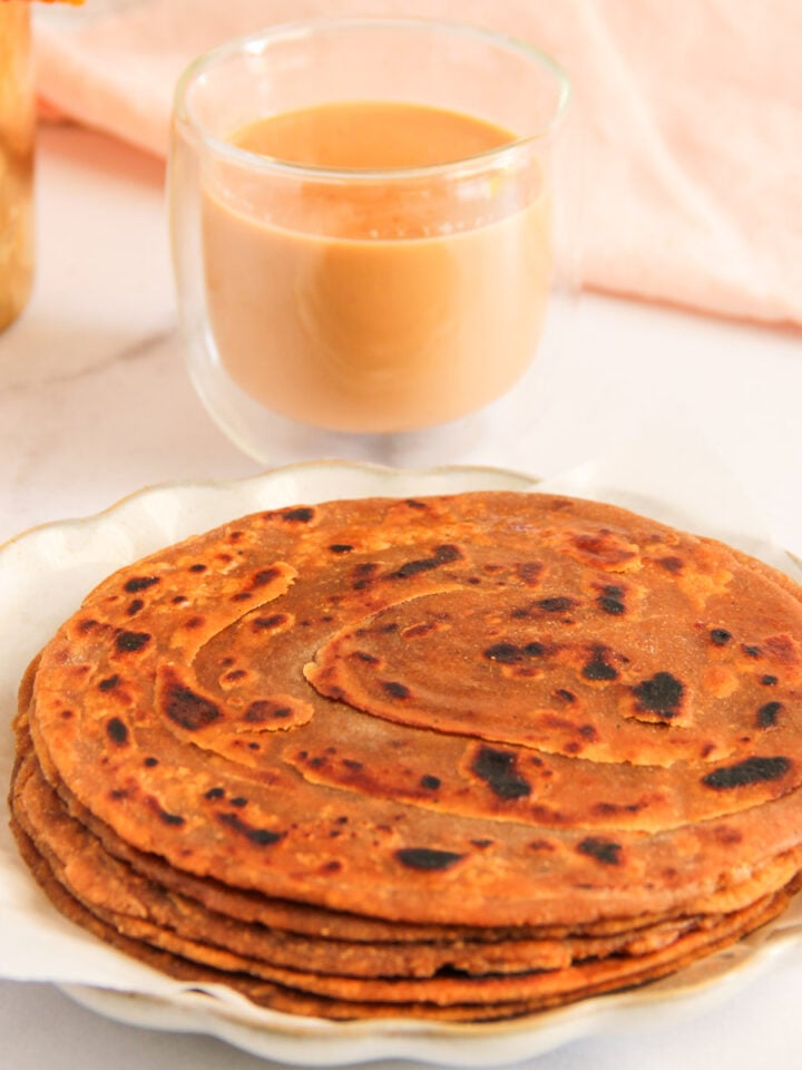 A plate of stacked parathas sits next to a glass of chai on a white surface with a pink cloth in the background.