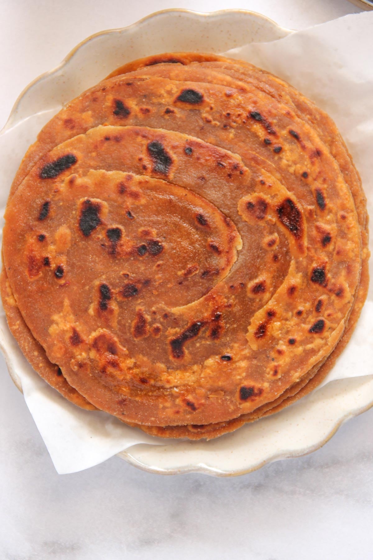 A plate with a stack of round, golden-brown parathas on white parchment paper.