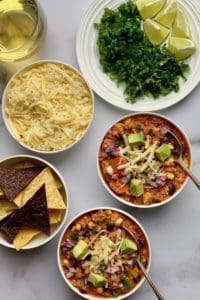 2 white bowls of buffalo chicken chili garnished with cheese and avocados, bowl of tortilla chips, cheese and a plate with chopped cilantro and scallions