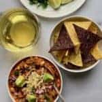 Bowl of Buffalo Chicken Chili garnished with cheese and diced avocados, with a bowl of tortilla chips, glass of drink and lime wedges in a white plate on the side