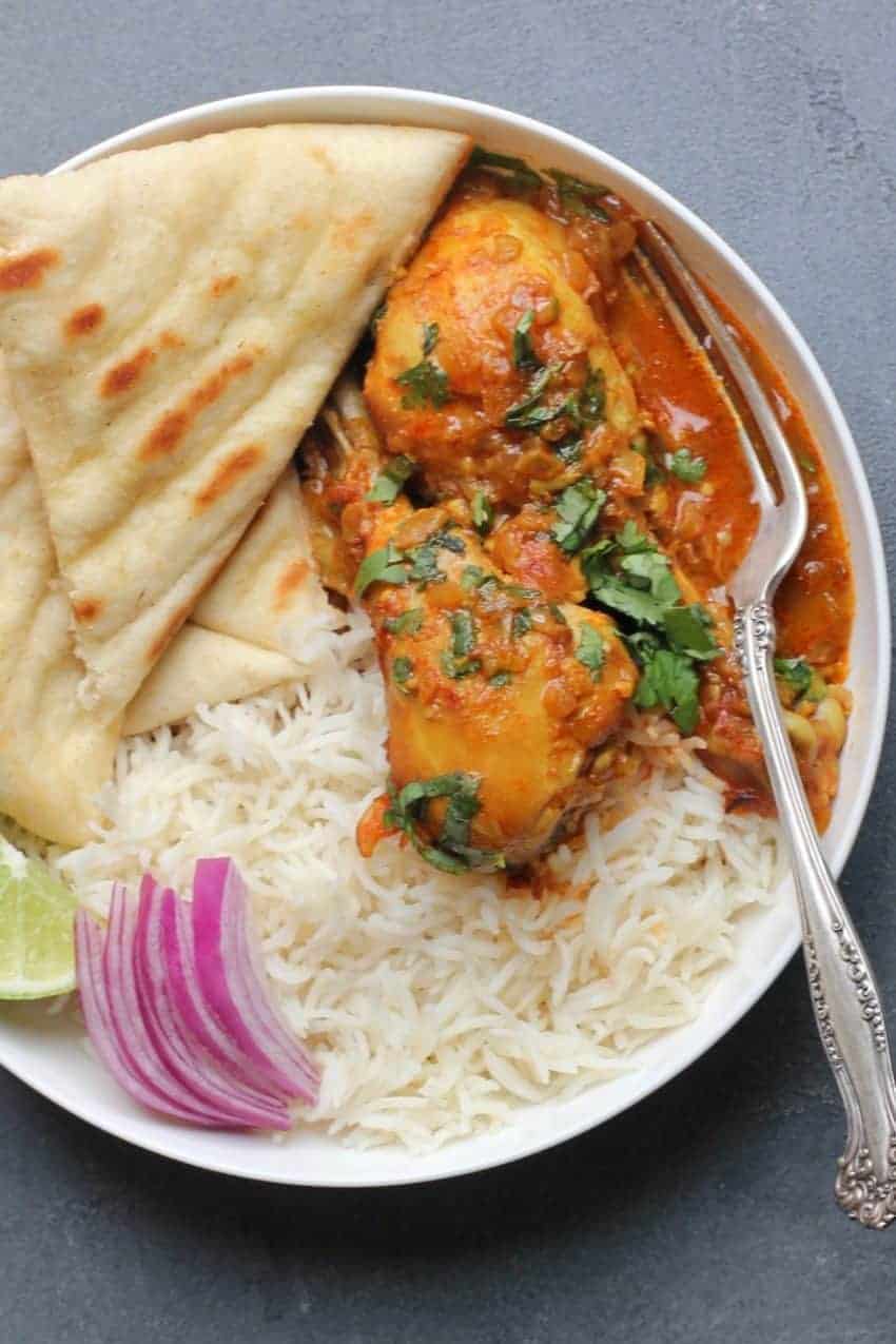 White bowl with Chicken korma curry served next to white rice and naan. Garnished with cilantro, sliced red onions and lime wedge.