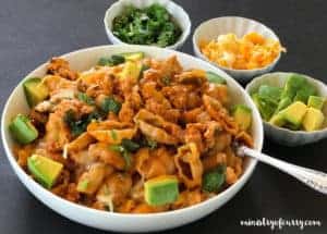 spicy taco pasta served in a white bowl, small bowls of cilantro, cheese and diced avocado on the side