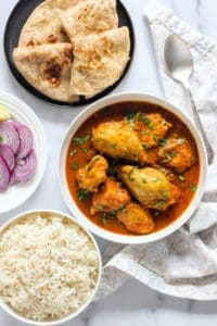 Chicken Curry in a white bowl with rice, chapatis and sliced onions on the side