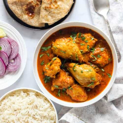Chicken Curry in a white bowl with rice, chapatis and sliced onions on the side