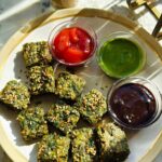 A plate of square pieces of kothimbir vadi garnished with sesame seeds, served with three dipping sauces: ketchup, green chutney, and tamarind chutney.