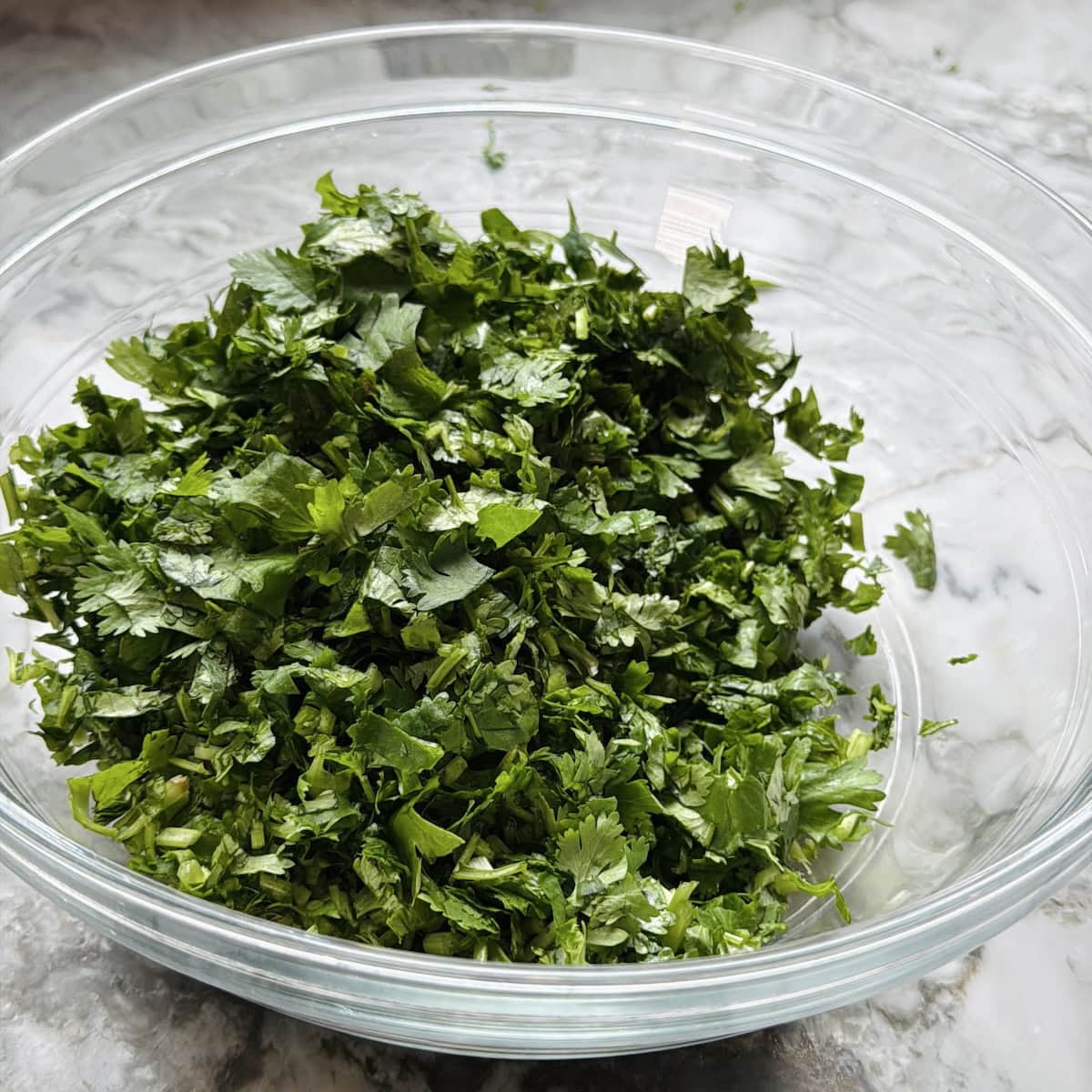 A clear glass bowl filled with freshly chopped cilantro, on a marble countertop.