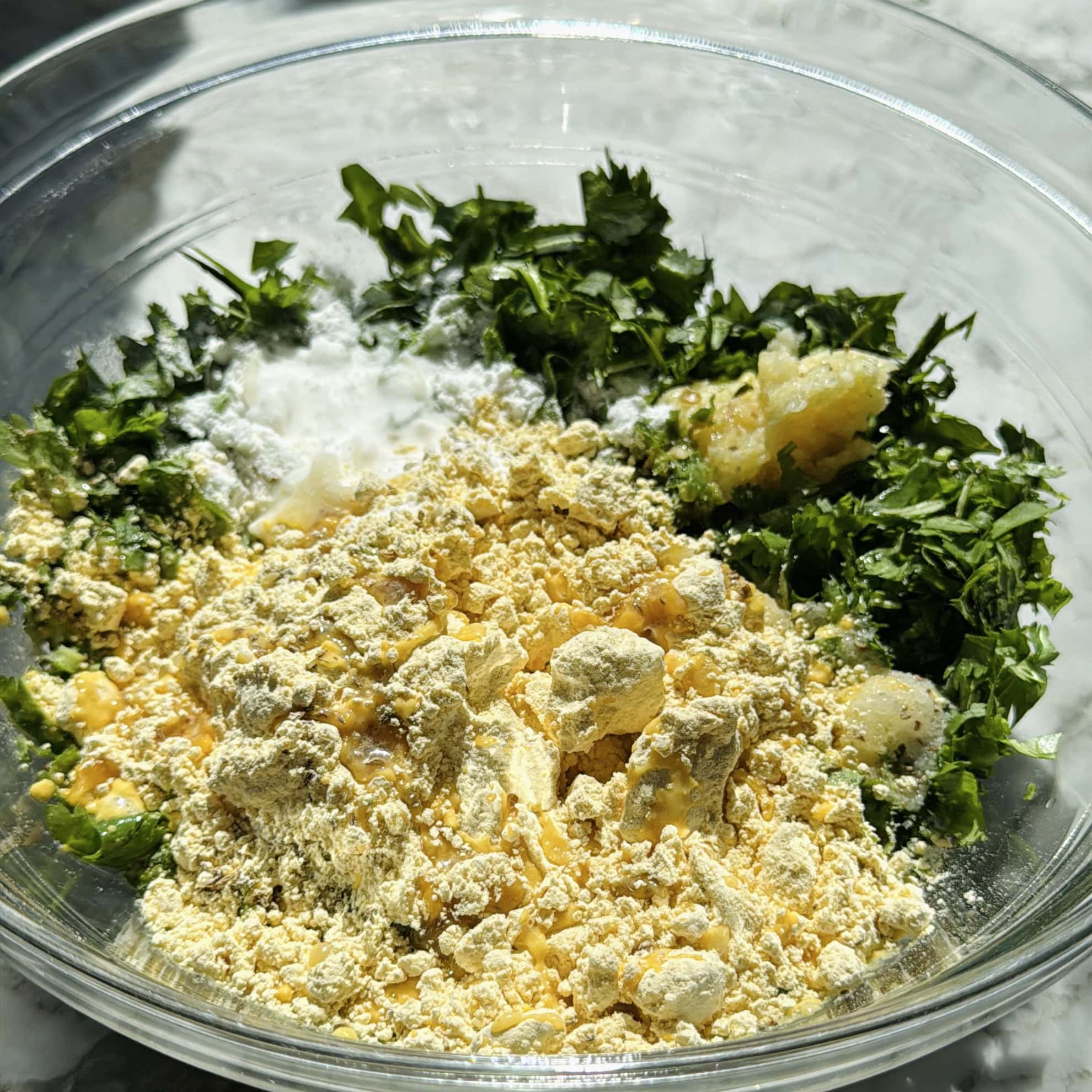 A clear bowl containing chickpea flour, chopped cilantro, garlic paste, and spices, ready to be mixed for cooking.