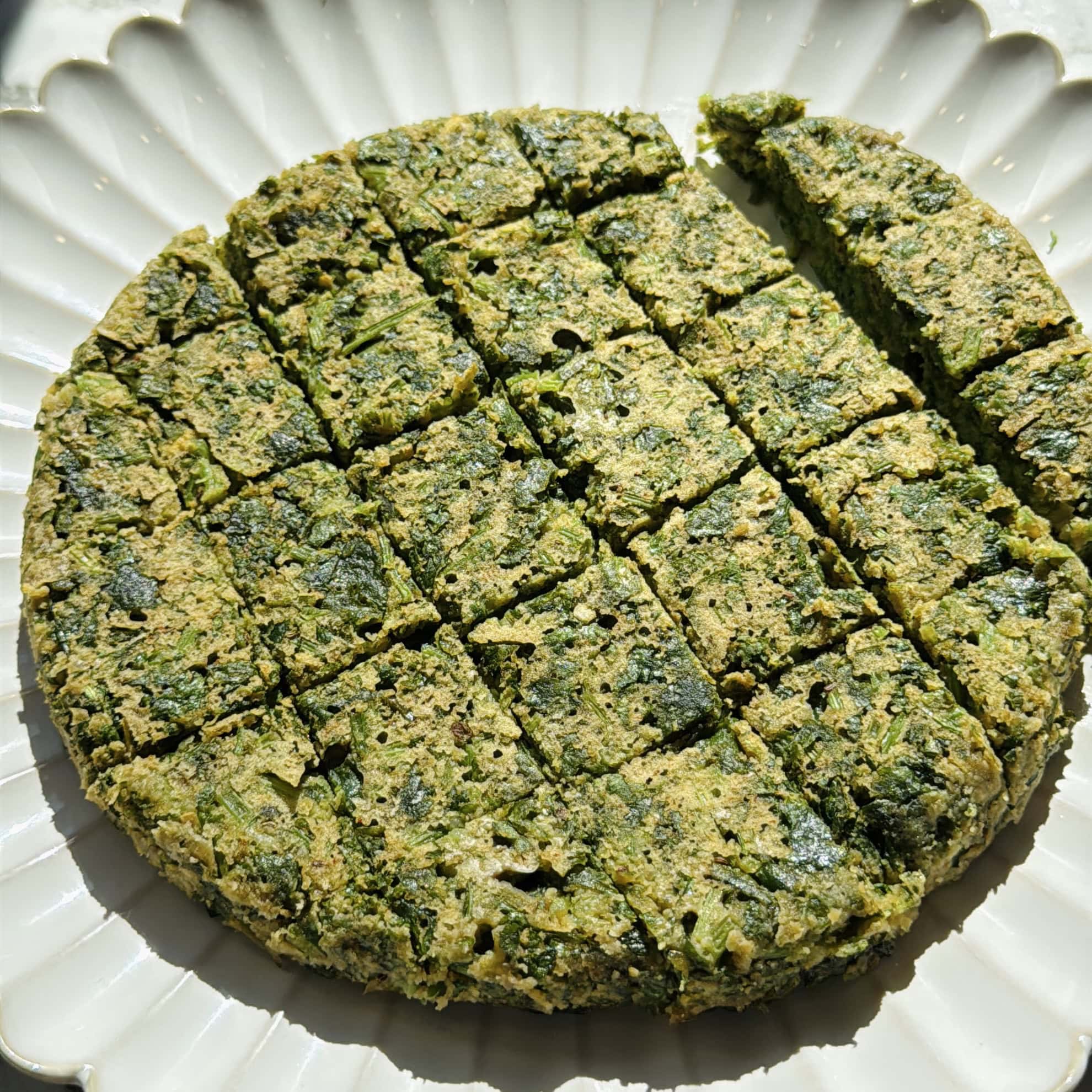 A round, kothimbir vadi cake cut into square pieces is arranged on a white, scalloped plate.
