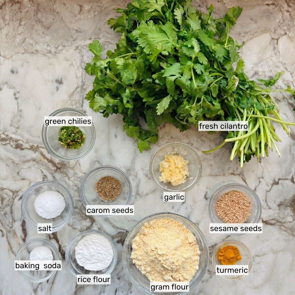 Ingredients to make Kothimbir Vadi: fresh cilantro, green chilies, carom seeds, garlic, sesame seeds, salt, baking soda, rice flour, gram flour, and turmeric.