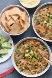 Two bowls with chicken tortilla soup, garnished with cubed avocados, cilantro and cheese. A plate on the left with lime wedges and chopped cilantro. Bowl of homemade baked tortilla chips.