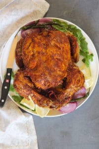 whole tandoori chicken served on a white platter with grilled red onion wedges, chilies, cilantro and lemon wedges.