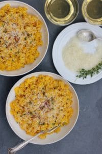 Saffron Risotto, garnished with parmesan cheese and saffron served in 2 white bowls. A plate of grated parmesan cheese on the side, with few srings of thyme and a spoon.