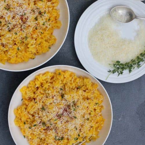 Saffron Risotto, garnished with parmesan cheese and saffron served in 2 white bowls. A plate of grated parmesan cheese on the side, with few srings of thyme and a spoon.