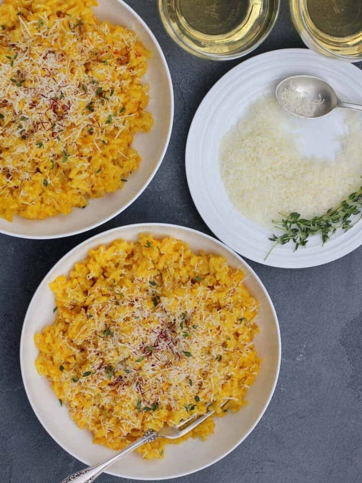 Saffron Risotto, garnished with parmesan cheese and saffron served in 2 white bowls. A plate of grated parmesan cheese on the side, with few srings of thyme and a spoon.