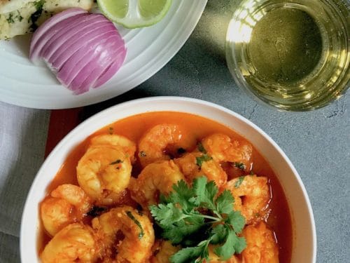 Shrimp curry in a white serving bowl, a white plate with cut up naan, sliced red onions and lemon wedges. Sparking drink in 2 cups on the side.