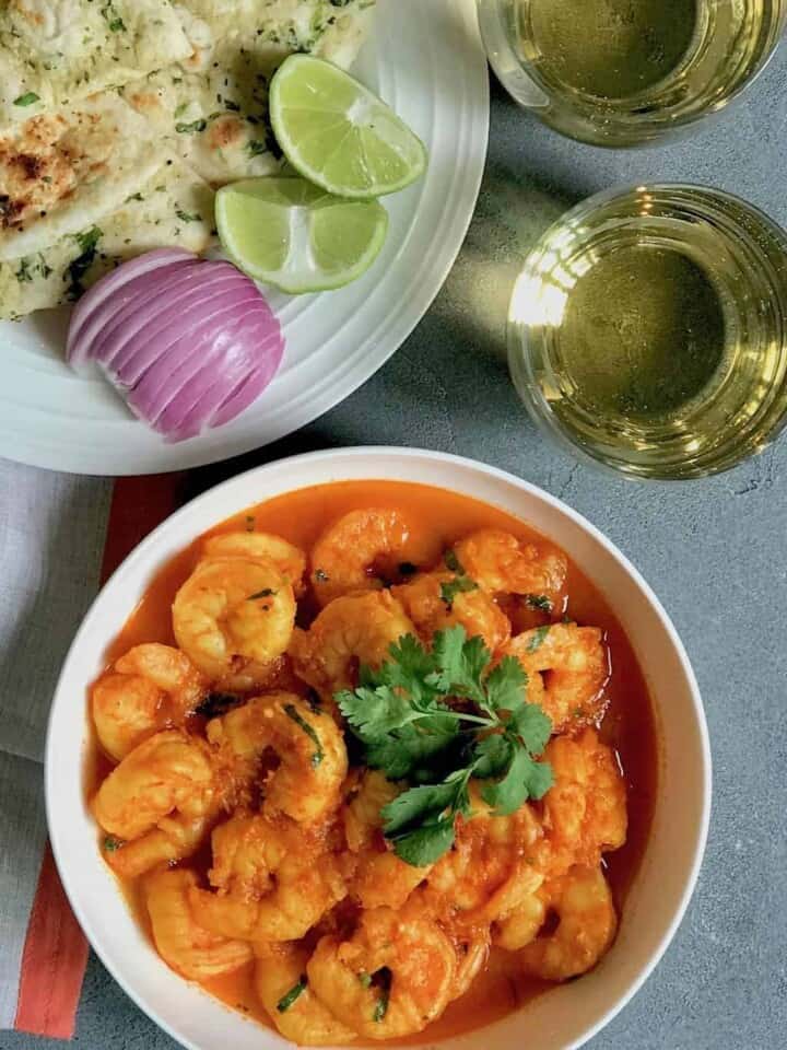 Shrimp curry in a white serving bowl, a white plate with cut up naan, sliced red onions and lemon wedges. Sparking drink in 2 cups on the side.