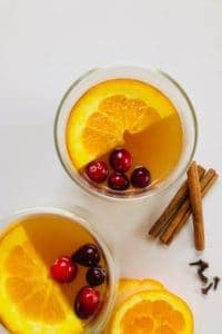2 cups with spiced apple cider, a wedge of orange and 4 cranberries floating on top. Orange slices, cinnamon sticks and cloves next to the cups on the table.