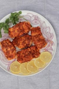 Tandoori Salmon fillets in a white platter surrounded by sliced lemons, sliced onions and cilantro