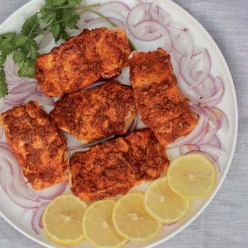 Tandoori Salmon fillets in a white platter surrounded by sliced lemons, sliced onions and cilantro