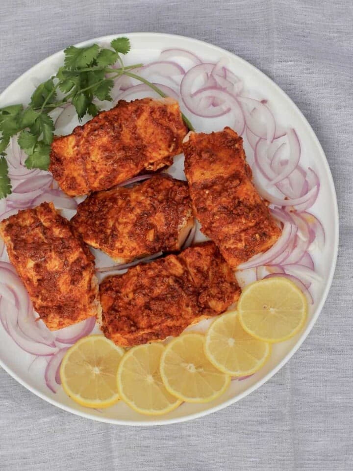 Tandoori Salmon fillets in a white platter surrounded by sliced lemons, sliced onions and cilantro
