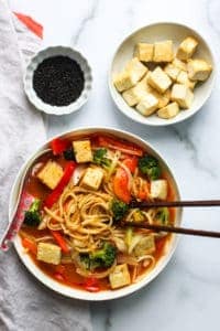 coconut curry noodle soup in a white bowl with chopsticks on one side and a soup soup on the other side immersed in the soup. A small bowl of stir fried tofu and black sesame seeds on the side
