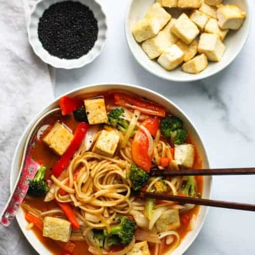 coconut curry noodle soup in a white bowl with chopsticks on one side and a soup soup on the other side immersed in the soup. A small bowl of stir fried tofu and black sesame seeds on the side