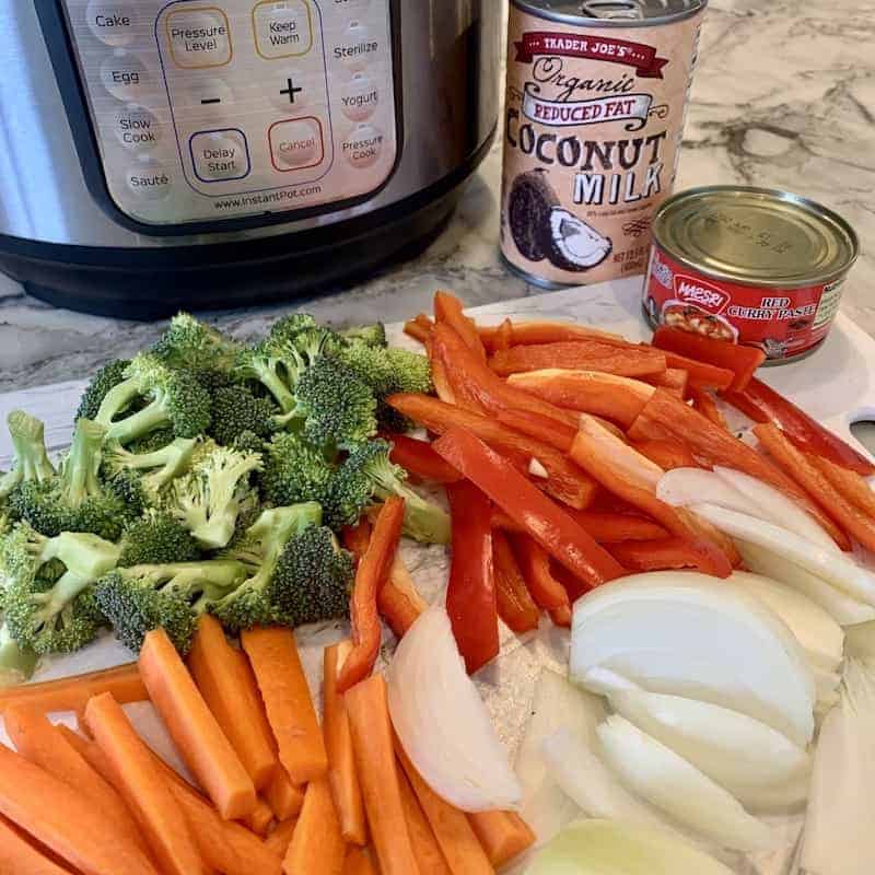 Carrots, broccoli, onions and red peppers cut on a cutting board with a can of coconut milk, red curry pasta on the side.