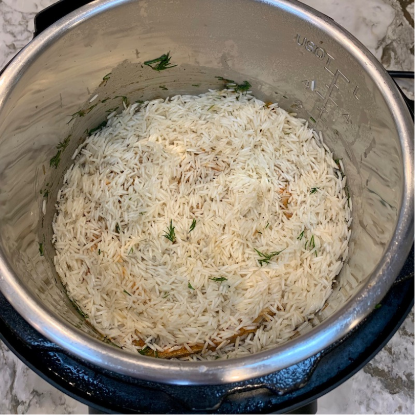 Uncooked basmati rice with some herbs inside a stainless steel Instant Pot on a marble countertop.