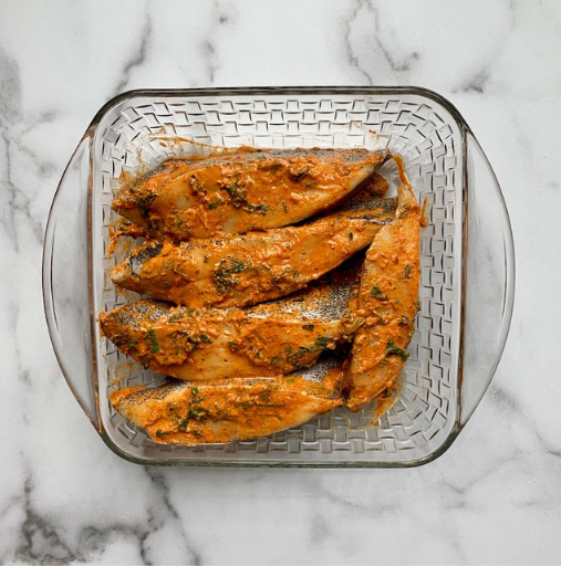 Glass dish containing six marinated fish fillets with herbs and spices, placed on a white marble surface.