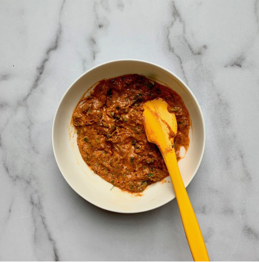 A white bowl with a yellow spatula contains a chunky, reddish-brown paste with visible herbs, placed on a light grey marble surface.